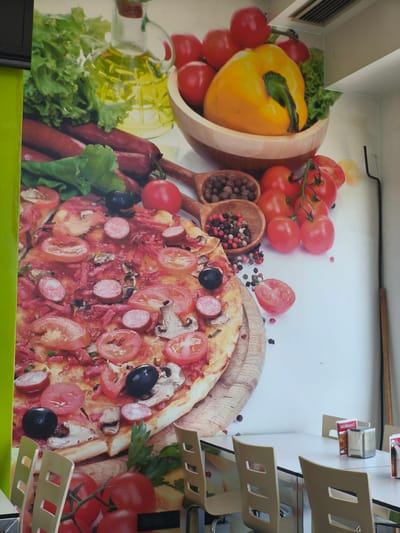 The image presents the interior of a casual dining establishment, likely a pizzeria or restaurant, in Moaña, Spain. The main feature is a large, vibrant wall mural depicting various fresh ingredients and a cooked pizza.

The mural's upper section showcases fresh green lettuce, a glass bottle of olive oil, a wooden bowl filled with bright red tomatoes and a large yellow bell pepper, and dark red sausages. The central part of the mural prominently features a large, generously topped pizza with sliced tomatoes, black olives, mushrooms, and round sausage or pepperoni slices, resting on a wooden cutting board. Around the pizza, wooden spoons hold black and mixed peppercorns, and fresh red tomatoes on the vine are scattered across the white background.

In the foreground, several light-colored dining tables with metal frames are set up, accompanied by light brown chairs featuring distinctive square cutouts in their backrests. On one table, a silver napkin dispenser and a red menu holder are visible. The menu holder clearly displays the Spanish word "¡NOVEDADES!" (News! / Novelties!). The lighting is bright, suggesting daytime conditions. A green wall or pillar is visible on the left side of the frame, and a ceiling vent is present in the top right. No people are visible, indicating a quiet moment in the restaurant.