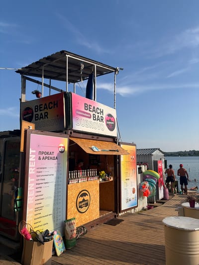 This media file depicts a lively beach bar scene on a sunny day. The main subjects are the "BEACH BAR DUBROVO" structures, likely shipping containers repurposed as kiosks, adorned with prominent signage. One sign, featuring a pink circle with a wave and the text "DUBROVO beach", is particularly eye-catching. Adjacent to it, another sign reads "BEACH BAR DUBROVO" in large, bold lettering.
Below the bar, a menu board is displayed with prices for various rentals and services in Cyrillic, including "катамаран" (catamaran), "лодка" (boat), "сап-борд" (SUP board), and "шезлонг" (sun lounger). The menu is divided into "будни" (weekdays) and "выходные" (weekends), indicating a resort or recreational area.
In the background, the calm water of a lake or river is visible, with a few people swimming or relaxing by the water's edge. Some individuals, seemingly staff or patrons, are standing on a wooden deck. Inflatable rings are piled up, suggesting water activities. The overall setting is a waterfront leisure area, likely a popular spot for summer recreation.
The bright sunlight and clear blue sky suggest it's daytime, probably mid-afternoon, given the angle of the shadows. The atmosphere appears relaxed and cheerful, typical of a beachside establishment. A red Coca-Cola cooler is visible, adding to the commercial aspect of the scene. The image is taken from a low angle, emphasizing the height of the bar structures and the expansive sky.