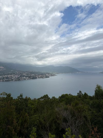 The image presents an elevated, panoramic view of what appears to be a coastal urban area situated on a narrow strip of land bordered by water on both sides. The location bears a resemblance to a coastal city, potentially Provodina, Montenegro, but lacks the concrete details to make an explicit claim.

To the right of the frame is dense foliage, suggesting a vantage point from a wooded hillside or mountain. Below, the urban area is densely populated with buildings, likely residential or commercial. The city extends along the thin peninsula, giving the impression of an elongated settlement.

The water on either side of the urban area appears calm. In the distance, more land masses are visible, suggesting either islands or a continuation of the coastline.

The sky is heavily overcast, with thick cloud cover dominating the upper portion of the image, hinting at potential unfavorable weather.