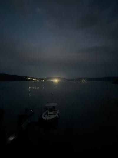 The image depicts a dark, atmospheric scene of a body of water at night. In the foreground, a small white boat with a canopy is moored on the right. To the left and slightly behind it, another darker boat is partially visible, with some lights illuminating its interior. The water is calm, reflecting the distant lights of a town or city along the shoreline in the background. The shoreline itself appears to be a hilly or mountainous landscape, with scattered lights indicating human habitation. The sky is overcast, with dark clouds obscuring any stars or moon. The overall impression is one of a quiet, somewhat eerie night scene. The image has a grainy texture, suggesting it was taken in low light conditions. There are no people visible in the image. The specific location of Zvanika, Bulgaria is not evident from the visual information provided in the image.