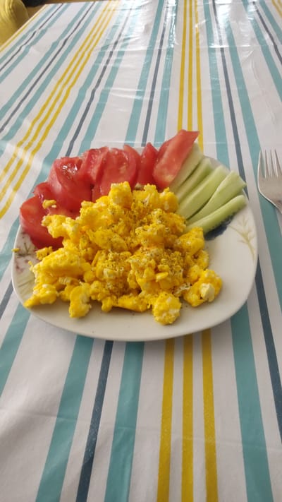A close-up, top-down shot showcases a plate of food on a striped tablecloth. The plate contains scrambled eggs seasoned with pepper, sliced tomatoes, and cucumber sticks. The tablecloth features alternating blue, yellow, and white stripes. A silver fork is partially visible on the right side of the frame. The location is identified as Kurtovo Konare, Bulgaria.