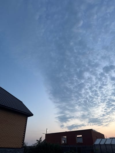 The image shows a partially constructed building and a finished house with a dark tiled roof. The sky is predominantly blue with a band of patchy, textured white clouds stretching across the upper right portion of the frame. The lighting suggests it is either early morning or late evening, with a subtle warm glow on the horizon. In the foreground, the side of a yellow brick house is visible, with a dark roofline. To the right, the brick structure of an unfinished building with open window frames is present, as well as a greenhouse-like structure with a white tarp. A wooden fence runs along the bottom of the image. Power lines and a utility pole are visible in the middle distance. The scene appears to be in a semi-rural or suburban area, consistent with a village or settlement in Russia.