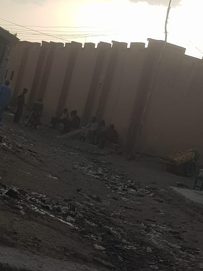 The image depicts a scene in what appears to be a narrow alley in Dutsinma A, Nigeria. A row of people are lined up against a tall, segmented wall. The ground is uneven and covered with debris. In the background, stacked yellow objects are visible near the top of the wall. The lighting suggests it is either early morning or late afternoon.