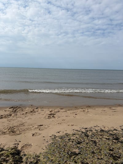 The image depicts a tranquil beach scene at Prestatyn, United Kingdom. The shot is oriented vertically, with the sky occupying the left half and the beach and sea on the right. The sky is filled with scattered white clouds against a pale blue backdrop, suggesting a bright but possibly breezy day.

The sea is calm, with gentle waves lapping at the shore. A thin line of white foam marks the waterline as it recedes from the sand. The beach itself is composed of light brown sand, marked with numerous footprints, indicating recent human activity. Further to the right, the sand transitions into a denser area covered with seaweed and small pebbles, with patches of green algae.

The overall impression is one of a peaceful day at the coast, likely during daylight hours given the brightness. There is no visible text in the image. The scene evokes a sense of relaxation and the natural beauty of a seaside environment.