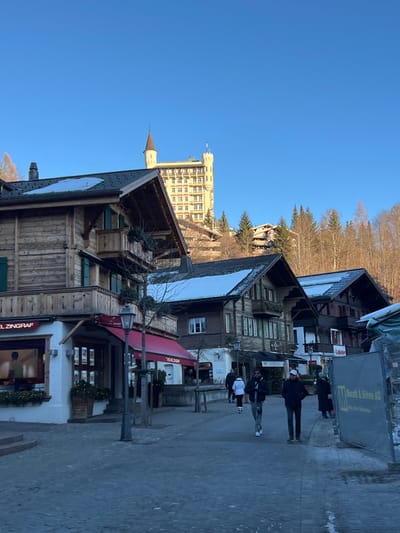The scene depicts a pedestrian street in Gstaad, Switzerland, with the iconic Gstaad Palace hotel visible on a prominent hillside in the background. The environment is bright, illuminated by strong sunlight under a clear blue sky, indicating a winter day due to snow patches on building roofs and some bare trees.

In the immediate foreground, a paved street or walkway occupies the lower half of the frame. On the left, a traditional multi-story Swiss chalet-style building with a rough-hewn wooden facade, green shutters, and a balcony on its upper level features a ground-floor commercial establishment labeled "HOTEL ZINGRAF." This shop has large display windows and a red awning. Adjacent to it, another building, possibly connected, also features a red awning and the partial text "THE ZINGRAF." A dark, ornate street lamp post stands in front of these buildings. Further down the street, several other chalet-style buildings with dark wooden facades, balconies, and pitched roofs are partially covered with snow.

On the paved street, approximately six to eight individuals are present. Three people are more distinctly visible in the mid-ground, walking. A person in a white jacket walks away from the camera on the left side of the path. Two men walk towards the camera in the center; one wears a dark jacket and light pants, the other a dark jacket and a white baseball cap. All individuals are dressed in winter attire. On the far right, a large, rectangular construction barrier or sign with "M. Buratti & Söhne AG" partially visible is present.

Dominating the background and situated on a heavily forested hillside, the Gstaad Palace hotel stands prominently. It is a large, light-colored, multi-story building characterized by numerous windows arranged in a grid pattern and distinctive pointed turrets, resembling a castle. Several other smaller chalet-style buildings are nestled among the dense coniferous trees on the hillside below the main hotel structure.