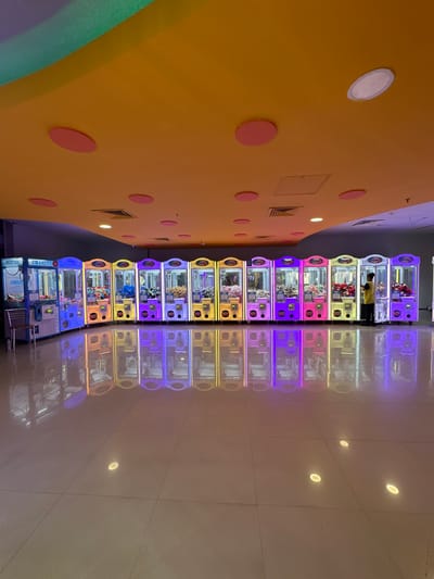 The image depicts a row of claw machines lining a wall in a hallway, likely within a shopping mall or arcade, based on the polished floor and wall design. The machines are colorful, with each seemingly having a different color scheme and filled with various prizes, mostly plush toys. A person, likely an employee, is standing between two of the claw machines. The scene appears to be in Kalyan-Dombivli, India, given the instructions. The wall beside the claw machines is painted yellow and adorned with pink circles. The floor creates a reflection of the claw machines. The lighting is bright, with the machines lit internally. The ceiling has recessed lighting.