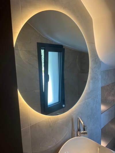 The image depicts a modern bathroom with a large, circular mirror illuminated by a warm LED light around its perimeter. The mirror is mounted on a wall tiled with large, grey marbled-effect tiles. Reflected in the mirror is a dark-framed window, partially open, revealing natural light and greenery outside. Below the mirror, a chrome faucet with a sleek, curved design is visible, positioned over a white ceramic sink. The overall aesthetic is contemporary and minimalist, suggesting a high-end bathroom design. The location is specified as Moni Community, Cyprus.