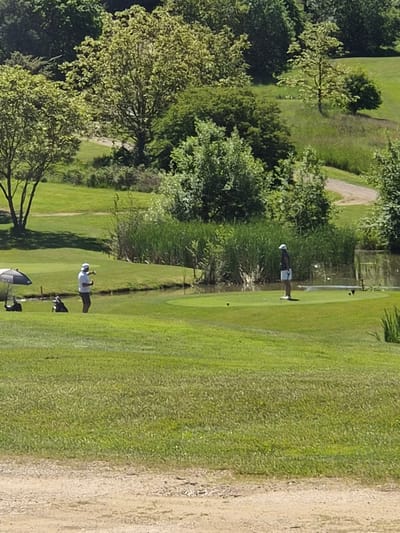 The image shows a golf course, likely located in Lenningen, Luxembourg. Two individuals are present: one is preparing to swing a golf club while another person is standing further away. The environment consists of grassy terrain, trees, and what appears to be a sandy area to the right of the frame. Golfing equipment, including bags and clubs, are visible next to one of the people.