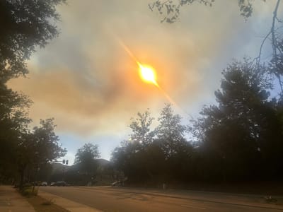 The photograph depicts a street scene in Oak Park, California, United States, under smoky conditions. The sun is visible as a bright, blurred orb, with prominent flares caused by atmospheric particles. The sky is hazy, with a mix of blue and orange tones, suggesting smoke or smog. Trees line both sides of the street, their foliage silhouetted against the bright sky. A road with two lanes is visible, with a sidewalk on the left side. Several cars are parked or moving in the distance. Traffic lights are also visible. The lighting is affected by the smoky atmosphere, creating strong shadows and reducing overall visibility.
