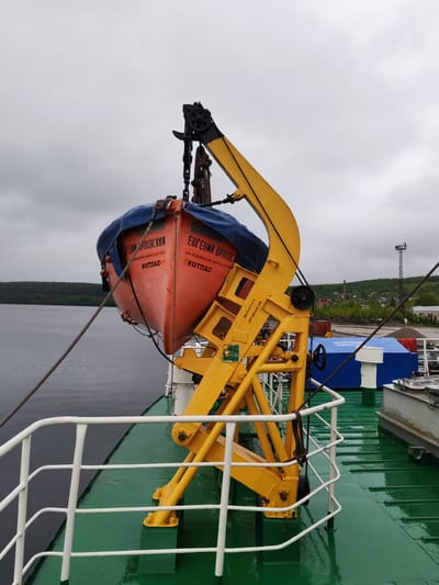 The image captures a bright orange lifeboat suspended from a yellow davit on the port side of a ship, against an overcast sky. The ship's green deck appears wet and reflective, suggesting recent rain or high humidity. This scene is likely set on a large body of water, possibly Lake Onega near Medvezhyegorsk, Russia, given the background of dense green forest covering hills.

The lifeboat is partially covered with a blue tarpaulin and features prominent Cyrillic text. On its bow, the name "ЕВГЕНИЙ ОРЛОВСКИЙ" (Yevgeny Orlovsky) is clearly visible, along with "АО РЫБИНСКОЕ ПАРОХОДСТВО" (JSC Rybinsk Shipping Company) and "КОТЛАС" (Kotlas), which likely identify the lifeboat's name, owner, and homeport, respectively. The yellow davit itself has codes "ИСП.04.24." (presumably "Inspection 04.24.") and "ПРОВ.04.26." (likely "Verification 04.26."), possibly indicating maintenance or testing dates. Further down the ship's deck, a blue cover bears the text "АВАРИЙНЫЙ ИНВЕНТАР" (Emergency inventory).

The davit structure is robust, equipped with black pulleys, chains, and thick ropes that secure the lifeboat. A white metal railing runs along the edge of the ship's deck. In the distance, beyond the calm water, a forested shoreline stretches, with a glimpse of an industrial or port area on the right. This includes a tall metal communication tower, what appears to be an old brick factory chimney, and other buildings. The overcast sky indicates a cloudy daytime, providing soft, diffused lighting to the tranquil scene.