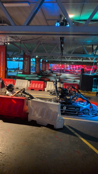 This is an indoor go-kart track at Grizzly.se in Östersund, Sweden. A child wearing a helmet is seated in a go-kart, holding the steering wheel. The go-kart is positioned on the track, which is delineated by red and white barriers. Several other go-karts are visible in the background, along with Coca-Cola branded banners and checkered flag motifs on the walls. The track is illuminated by colored lights, casting blue and green hues on the ceiling and track surface. The overall environment suggests an active recreational setting.