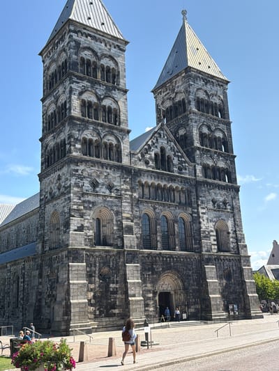 This image captures the Lund Cathedral, a prominent landmark in Lund, Sweden. The cathedral is constructed from dark, weathered stone and features two imposing bell towers. The right tower is topped with a conical, metallic spire, while the left tower has a more angular, segmented roof. Arched windows and decorative stone carvings adorn the facade.
In the foreground, a woman with dark curly hair and a backpack walks away from the camera on a paved walkway. She is wearing a light-colored denim skirt and a white top. To the left, a planter filled with vibrant pink and red flowers sits near a bench where two people are seated. Several other individuals are visible near the entrance of the cathedral. The sky is a clear, bright blue with a few wispy clouds. The overall scene depicts a sunny day in a historic European city.