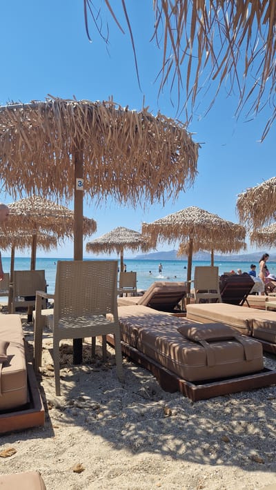 The image captures a view of a beach in Artemida Municipal Unit, Greece, under a clear blue sky. Several thatched umbrellas provide shade over rows of empty, beige lounge chairs on a sandy beach. In the background, the calm sea is visible with a few people swimming and wading. The shoreline is dotted with more umbrellas and lounge chairs. The overall atmosphere is relaxed and sunny, typical of a beach resort.
