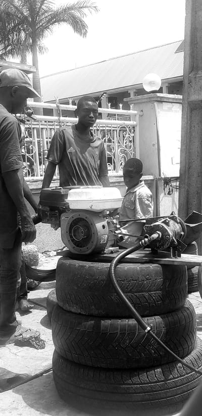 In Kinkinau, Nigeria, three individuals are gathered around a generator unit precariously stacked on a base of tires. An adult male, wearing a cap and dark t-shirt, stands on the left, his hands near the generator's frame. Beside him, another adult male, in a dark t-shirt with an Adidas logo, looks towards the generator. To the right, a young boy, dressed in a light-colored collared shirt, observes the machine. The generator itself is a central focus, with its engine and fuel tank visible. A black hose extends from the generator and connects to another piece of equipment on the right. The scene unfolds outdoors, with a building and a palm tree visible in the background, suggesting a commercial or residential area. The ground is paved, and other discarded tires are present.