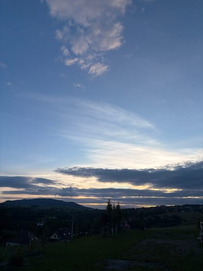 The image captures a twilight scene in Stara Wieś, Poland. The sky is a gradient of deep blue at the top, transitioning to lighter blues and streaks of white and yellow clouds closer to the horizon. A band of darker, textured clouds is prominent just above the landscape.

The landscape below features rolling hills covered in dark green foliage. Several houses with pitched roofs are visible nestled amongst the trees and on the hillsides. In the foreground, a grassy area transitions to a darker, rougher texture, possibly gravel or soil. A few trees, including two tall, thin evergreens, stand in the middle ground. A distant, dark silhouette of a mountain range or large hill forms the backdrop against the colorful sky. The overall impression is one of a peaceful rural evening.