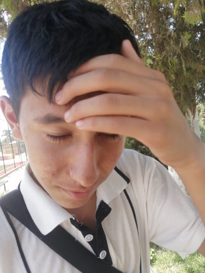 A young man, likely in his late teens or early twenties, is captured in a close-up shot outdoors. He has short, dark hair and his right hand is placed on his forehead, partially obscuring his face. His eyes are closed, and his expression suggests contemplation or discomfort. He is wearing a white polo shirt with black trim around the collar and down the placket, featuring visible white buttons. The background reveals a hint of trees with light green foliage, suggesting a park or natural setting within a city. In the distance, a blurred structure with a fence and what appears to be a track or field can be observed, possibly indicating a sports facility or educational institution. The lighting suggests it is daytime. The image does not contain any specific landmarks or elements that definitively place it in Sapzipaya, Uzbekistan.