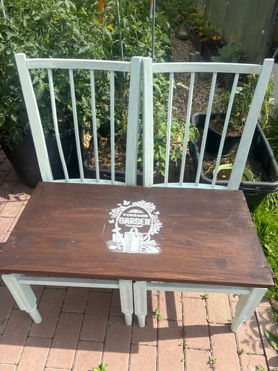 The image shows a wooden bench with a light blue painted frame and a dark brown seat. The seat is decorated with a white stencil that depicts a watering can, some plants, and butterflies, with the word "GARDEN" in the center. The bench is placed outdoors on a brick patio, with a garden in the background. The garden appears to be a vegetable garden with tall green plants, some of which are supported by stakes. There are also some black plastic planters and a wheelbarrow visible in the background. The lighting suggests it is a sunny day. The location is described as Lotosh SNT (Maryino), Russia.