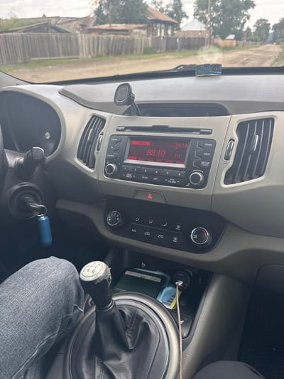 The image shows the interior of a manual transmission car. The dashboard displays a car radio tuned to FM/AM, showing the time as 18:14 and a station frequency of 88.10. A mobile phone charger is plugged into the car's power outlet, with a phone cable connected. The driver's leg, clad in blue jeans, is visible next to the gear stick. Outside the car, through the windshield, a wooden fence and houses line a dirt road, suggesting a rural setting in Yabogan, Russia. The sky is overcast.
