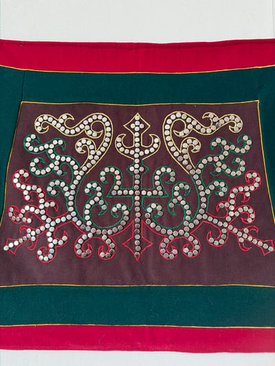 The image shows a close-up of a richly decorated piece of fabric, possibly a textile or part of an garment or tapestry. The main decorative element is an intricate embroidered pattern on a dark brown background. This embroidery features swirling, organic shapes outlined with small, metallic studs. The outlines themselves are done in gold, green, and red thread, with the metallic studs following these lines closely, creating a textured and shimmering effect. Above and below this central brown panel are horizontal bands of fabric in deep green and red, separated by thin gold piping. The overall impression is one of traditional craftsmanship and vibrant color. There are no people, animals, or discernible environmental features in the image. The location in Мяндиги, Russia is not depicted or suggested by the content of the image itself.