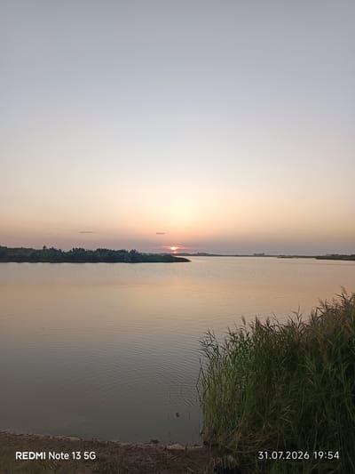A landscape photograph captures a serene sunset over a body of water. The sky transitions from a pale blue at the top to warm hues of orange and pink near the horizon, where the sun is partially obscured by a distant landmass. The water's surface is calm, reflecting the soft colors of the sky and creating gentle ripples. A line of trees and vegetation runs along the far bank of the water, creating a natural silhouette against the twilight sky. In the foreground, tall reeds and grasses grow along the water's edge. The bottom of the image displays text overlay: "REDMI Note 13 5G" on the left and "31.07.2026 19:54" on the right. The scene appears to be a natural setting within or near the city of Kabanbai Batyr, Kazakhstan, suggesting a peaceful evening by the water.