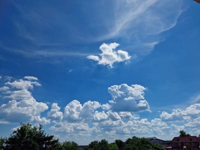This is a landscape photograph taken from a high vantage point, likely an upper floor or rooftop, in Krosno Odrzańskie, Poland. The image is dominated by a bright blue sky filled with various types of clouds. In the upper portion of the sky, thin, wispy cirrus clouds form delicate streaks, suggesting high-altitude winds. Below these, larger cumulus clouds are present, ranging from small, isolated puffs to larger, more developed formations with distinct shadows. The foreground features a line of dense green trees, including what appears to be an evergreen tree on the left. In the distance, visible above the trees, are parts of buildings. On the right side of the frame, a red-tiled roof of a building is clearly visible, suggesting a residential or commercial area. Further to the left, a larger, more modern building with a light-colored, curved roof structure is partially visible, possibly a public or industrial building. There are no people or animals clearly discernible in the image. The overall impression is that of a sunny day with dynamic cloud formations over a mixed urban and natural landscape.
