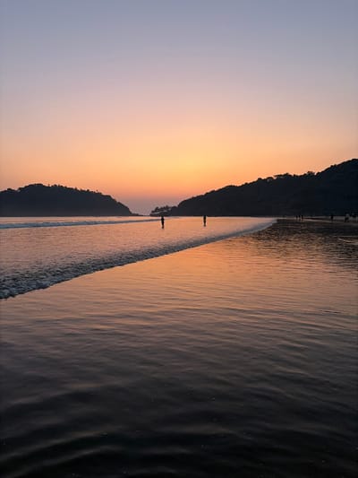 The image presents Kolamb Beach in Kolamb, India, during twilight. The sky exhibits a gradient, transitioning from light purple at the top to orange and yellow at the horizon. Two dark, tree-covered headlands frame a central bay. The ocean surface reflects the sky's colors, with gentle waves breaking in the foreground. Two silhouetted human figures stand in the shallow water near the image's center, while several smaller, silhouetted figures are discernible along the distant right shoreline.