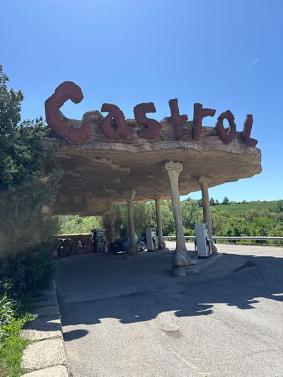 The image depicts a large, brown, sign with red lettering that reads "Castello" in Belomortsi, Bulgaria. The sign is supported by bone-like pillars and is located at a gas station. There are trees and foliage at the base of the sign. Cars and gas pumps are visible in the background. The sky is blue and clear. There is a contrail in the sky. The ground is asphalt and there are shadows cast upon it.
