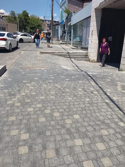 In an outdoor urban setting, a woman with red hair wearing a purple shirt and dark pants is walking towards the right side of the frame. She is positioned near a large dark entryway. To her left, further down a sidewalk with cobblestone paving, a woman in a black tank top and blue jeans is walking towards the left, followed by a man in a white shirt and dark shorts carrying a backpack. Several white cars are parked on the street on the left side of the image, with a white car in the foreground on the left. Buildings line the street, with shops visible in the background on the left. The sky is blue with some clouds. A shadow of a wire stretches across the cobblestone pavement.