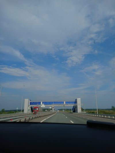 The image shows a view from inside a vehicle traveling on a highway in Kirovskoe S/P, Russia. A red truck is visible driving on the highway in the distance, approaching a large pedestrian overpass. The overpass has a blue and white structure spanning across the highway. The sky is partly cloudy with white clouds against a blue background. The environment appears to be rural, with green fields and trees visible on the sides of the highway. The foreground shows the dashboard of the vehicle.