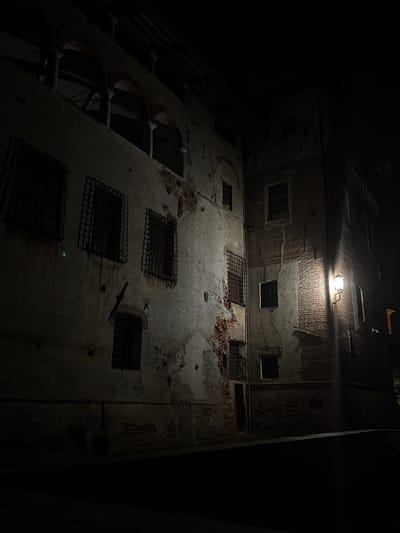 The image shows the exterior of a dimly lit, aged building at night in Lagnasco, Italy. The architecture is a mix of weathered plaster and exposed brickwork, with some sections showing signs of decay and damage. Numerous windows are visible, many of which are covered with heavy metal bars, suggesting a historical or fortified structure. An outdoor lamp with a warm glow illuminates a section of the brick wall, casting shadows and highlighting the texture of the building. The surrounding area is dark, with the street or pathway in the foreground obscured by shadow. The scene evokes a sense of history and perhaps abandonment or a secluded atmosphere.