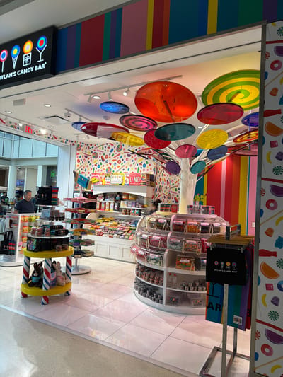 The image shows the interior of Dylan's Candy Bar, a candy store, at Charlotte Douglas International Airport in Charlotte, North Carolina. The store is brightly decorated with a colorful candy-themed interior. Large, circular, translucent candy-shaped decorations hang from the ceiling. The walls are covered in a vibrant pattern of candy images. Shelves stocked with various candies are visible. In the foreground, there is a display stand with candy and stuffed animals. A man stands in the middle ground looking towards the displays. The floor is tiled and reflective, showing some of the colorful decor. The overall impression is of a whimsical and fun environment.