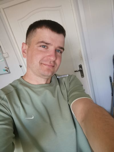 This is a selfie of a man with short dark hair and blue eyes. He is wearing a light green t-shirt with a white Nike logo on the left side of his chest and white trim on the sleeves. He is smiling and looking at the camera. The background appears to be indoors, with a white door and a door handle visible behind him. There is also a whiteboard with some writing and drawings on it to his left. The lighting suggests it is daytime. The man appears to be relaxed and in good spirits. A small white tag with black text is visible on the left side of his shirt near his shoulder. There is also some writing on the sleeve of his shirt. The overall impression is a casual and personal photograph.