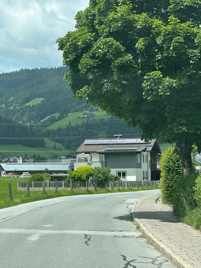 The image captures a rural scene in Reitdorf, Austria, featuring a paved road curving to the right, bordered by a brick sidewalk. A large, leafy green tree dominates the foreground on the right, casting a shadow onto the road. In the middle ground, a modern house with solar panels on its roof is visible, along with a wooden fence and some hay bales in a grassy field. Beyond the buildings, rolling green hills covered in dense forest rise up, with a few scattered houses dotting the landscape. The sky above is cloudy. The overall impression is one of a tranquil, picturesque Austrian village setting.