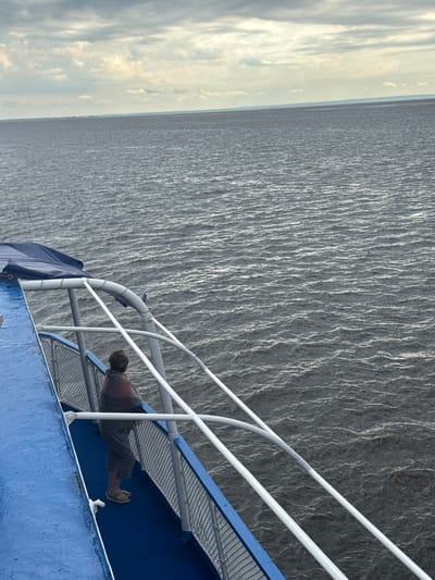 An individual stands on the blue deck of a boat, leaning against a white railing, looking out at a wide expanse of dark, rippling water. The individual wears a striped, draped garment over a brown shirt and shorts, and light-colored sandals. The boat's railing is secured by vertical supports and features a blue covering overhead. The water stretches to a distant, hazy horizon under a cloudy sky. The image captures a moment of quiet observation from the vessel, likely in or near Ikskoe Ust'e, Russia, given the context.