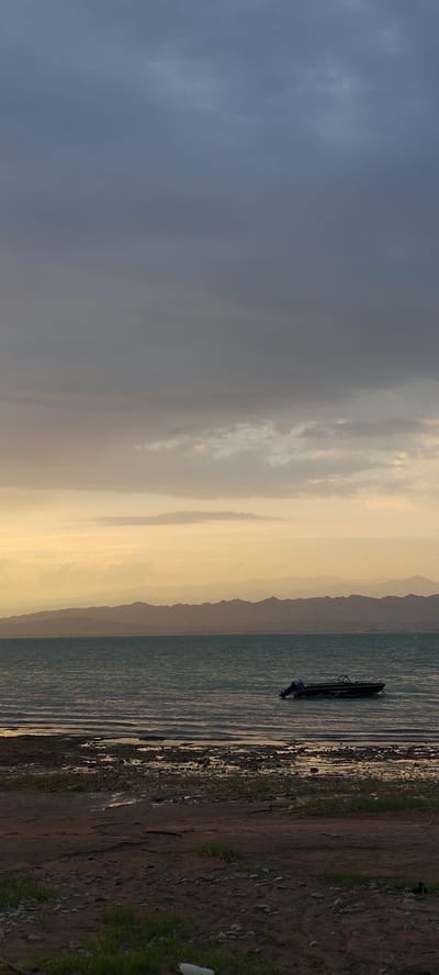 A lone boat with an outboard motor is anchored in the wavy waters of a large body of water, possibly a lake or reservoir, near a rocky, muddy shore with sparse vegetation. In the background, silhouetted mountain ranges stretch across the horizon, fading into a hazy sky. The sky transitions from a dark, cloudy expanse at the top to a band of warm, golden light near the horizon, suggesting either sunrise or sunset. The location is identified as Chigdalik, Tajikistan. There are no people visible in the scene.