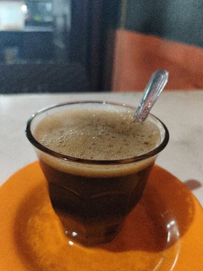 A high-angle, close-up photograph depicts a clear glass containing a dark, foamy beverage, likely coffee, situated on a bright orange circular saucer. A small, metallic spoon is inserted into the liquid, resting against the glass rim. The saucer and glass are positioned on a light-colored, granular surface. The background is out of focus, revealing blurred dark shapes on the left and a prominent reddish-orange surface on the right. The scene is illuminated by ambient light, suggesting an indoor setting within the city of Rheng, Indonesia. No individuals are present.