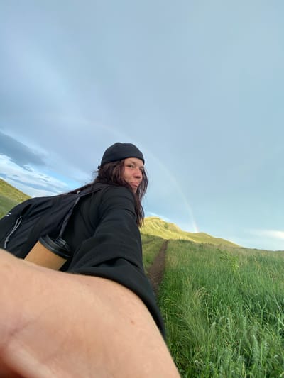 In a selfie taken outdoors, a person wearing a black beanie and black clothing looks back at the camera with their tongue sticking out. They have a backpack on their shoulder with a reusable coffee cup attached. The background shows a grassy hillside with a dirt path winding up it, and a faint rainbow in the pale blue sky. The location is not discernible from the image, and therefore cannot be identified as being in Donskoe, Russia.