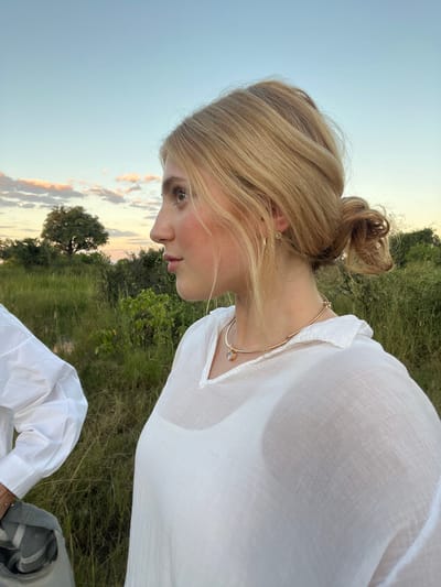 A young woman with blonde hair pulled back into a bun is shown in a low-angle, close-up shot, gazing downwards. She is wearing a sheer white button-down shirt and a delicate gold necklace. The background shows a grassy field with trees, and the sky is visible with a sunset. The location is Lobilos, Botswana. The subject is sitting.