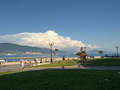 The image depicts a scenic waterfront promenade with a clear blue sky and dramatic, large cumulus clouds forming over distant mountains. In the foreground, a well-maintained grassy area is visible, dotted with small white flowers. A paved walkway runs parallel to the water, separating the grassy area from a concrete embankment.

Along the promenade, several people are casually strolling and sitting on benches. To the right of the image, a bronze sculpture of a woman riding a dolphin is prominently displayed. A purple electric scooter is parked in front of the sculpture. The sea stretches out beyond the embankment, with several ships and boats visible on the horizon. The distant shoreline is lined with buildings and industrial structures, suggesting a port or coastal city. The overall atmosphere is one of a pleasant, sunny day by the sea. The scene is likely located in Grushovaya Balka, Russia, given the context of the request.