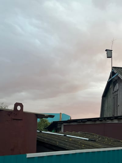 The image shows a cloudy sky over a cluster of buildings. In the foreground, a dark red structure with a large circular cutout is visible. To the right, a weathered wooden building with a pitched roof and a window stands tall. Atop this wooden building, a birdhouse is mounted on a pole, with a thin branch extending from it. Below these structures, other rooftops can be seen, including one covered in corrugated material and another with a teal-colored surface. Trees are present in the background. The scene appears to be in an outdoor, possibly rural or semi-urban, environment in Большой Кукмор, Russia, given the context. There are no people or discernible actions taking place. The overall atmosphere is subdued, likely due to the overcast sky.