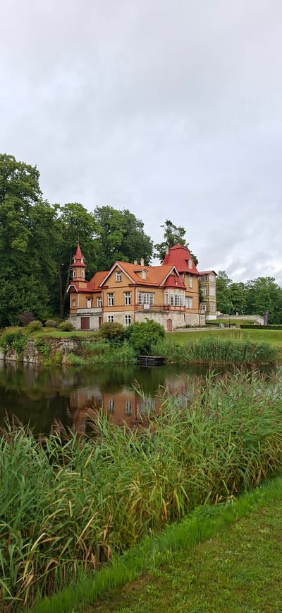 The image captures a serene waterside scene in Kuressaare, Estonia, with a historic building situated amidst lush greenery. The main subject is a multi-story, light-brown wooden building with a striking red roof, featuring intricate architectural details like dormer windows and turrets. The building is surrounded by mature trees, their dark green foliage contrasting with the building's warm tones.
A calm body of water, likely a canal or a small lake, runs alongside the building, reflecting its image and the surrounding vegetation. Tall, reedy grasses grow densely along the water's edge, partially obscuring the building's foundation. On the opposite bank, a neatly mowed grassy area slopes away, suggesting a park or garden setting. The sky is overcast, indicating a cloudy day, possibly in the late afternoon or early evening, given the soft, diffused light. There are no people visible in the image, contributing to the tranquil and undisturbed atmosphere of the location. The scene evokes a sense of peace and historical charm, typical of the Estonian coastline. No visible text is present in the image.