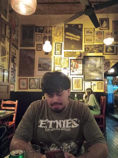 The image shows a man sitting at a table in a restaurant in Victoria, Argentina, located at latitude: -34.452, longitude: -58.54. The man is wearing a green t-shirt with the word “ETNIES” printed on it, black hair, and has a beard. He is looking down at a cell phone, with a green soda can and a glass of water on the table in front of him. There is another person, a woman with long dark hair, seated at a nearby table. The walls are decorated with pictures and posters. There are also several wooden tables and chairs in the restaurant. There is an overhead fan in the corner, with a light hanging from the ceiling. The restaurant is dimly lit, and there is a wooden door in the background.