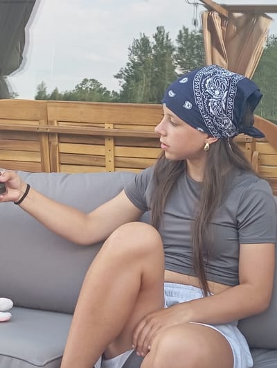 A young woman is seated on a grey couch, facing right, with her legs crossed. She wears a dark blue bandana on her head and a grey t-shirt. Her right arm is extended, holding a dark object, possibly a phone. She is wearing a gold earring in her visible ear and a black wristband on her left wrist. The background features a wooden structure with a tan canopy and a reflection of trees in a window. The scene appears to be an outdoor seating area, possibly on a patio or deck. There is no indication that this location is in Dybów Kolonia, Poland.