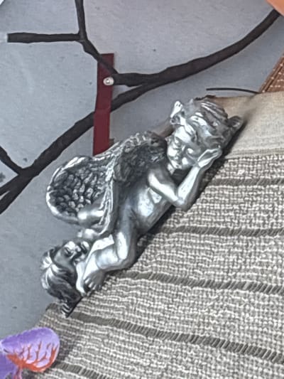 The media file captures a close-up, angled view of a metallic figurine of two cherubs, rendered in a silver-toned material. One cherub is positioned above the other, appearing to recline and rest its head on its hands, while the lower cherub's head is visible beneath the upper one's torso. Both figures have delicate wings and youthful features. The figurine rests on a textured, grey surface, possibly a wall or fabric, which has a woven pattern. A dark, branch-like element extends from the left side of the frame, with a thin, dark red horizontal bar visible beneath the figurine. In the lower right corner of the frame, a small portion of a purple and orange object, possibly a flower or decorative element, is visible. The lighting suggests an indoor setting, with no specific time of day or weather conditions indicated. No visible text is present in the media file. The location context of Osterode am Harz, Germany, is provided, but offers no direct visual cues within the image itself.