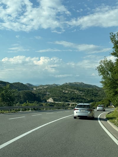 The image depicts a highway scene in the vicinity of Vrela, Montenegro, on a sunny day with scattered clouds. Two silver cars are traveling on a paved road with visible white lane markings. The car in the foreground, a silver station wagon, has a license plate that appears to read "BG 3D7A-TB". A second, smaller silver car is visible further ahead on the same road. The road is bordered by a metal guardrail and lush green vegetation. In the background, rolling hills covered in dense green trees and dotted with some scattered houses extend towards the horizon. The sky is a bright blue with fluffy white clouds. The overall impression is a scenic drive through a mountainous, vegetated region.