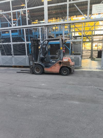 A bright, sunny day in Dimona, Israel, illuminates an industrial setting where a weathered orange and black Toyota forklift sits idle on an asphalt lot. The forklift, a robust industrial object, is the primary subject, its tines extended forward, suggesting recent or imminent use. Behind it, a large, metal-framed structure with wire mesh infills stands, likely part of a factory or storage facility. This structure houses stacked industrial materials, possibly tires or other large cylindrical objects, creating a sense of scale and industrial activity.
The background reveals more of the facility, with additional metal structures and what appears to be an open-air area with trees bathed in the warm glow of the sun. The time of day is clearly daytime, with strong sunlight casting shadows and highlighting the textures of the asphalt and metal.
A prominent sign is visible on the upper right portion of the metal structure. The text on the sign is in both Hebrew and English. The English portion reads: "LIQUID WASTE TREATMENT PLANT". This text provides crucial context about the location's purpose.
The overall scene conveys a sense of functional industrial space, designed for material handling and processing. The forklift's presence suggests ongoing operations, even though it is currently stationary. The bright sunlight and clear visibility point to favorable weather conditions. The scene is devoid of people, emphasizing the machinery and the infrastructure of the facility.