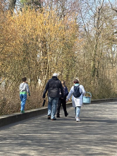 A daytime image in Bad Nauheim, Germany, depicts four individuals walking away from the camera on a paved asphalt path, flanked by a low concrete curb and dense vegetation, under a clear blue sky. A young boy, with light brown hair, wearing a grey long-sleeved shirt, blue trousers, and bright green shoes, walks on the concrete curb on the left. To his right, on the asphalt path, an adult male in a grey baseball cap, dark jacket, and blue jeans, holds a black cylindrical object in his right hand. Adjacent to him, a younger individual, dressed in dark attire, walks on the path. Furthest right, an adult female with reddish hair, wearing a light grey long coat, light blue jeans, white sneakers, and a dark blue backpack, carries a light blue plastic basket in her left hand. The asphalt path displays multiple elongated shadows, indicative of bright sunlight. The background consists primarily of bare deciduous trees and bushes, with one prominent bush in the mid-left section densely covered in light brown catkins.