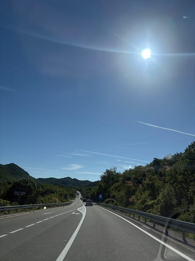 The image depicts a view from a vehicle moving along a winding road through a mountainous, forested landscape. The sun is bright and high in the sky, suggesting it is midday. The sky is a clear, deep blue with a few faint contrails from airplanes visible.
The road, paved with asphalt, has a white solid line marking the edge and dashed white lines indicating lanes. A metal guardrail runs along the side of the road, separating it from the steep, tree-covered slope. The trees are dense and lush, indicative of a warm, possibly summer season.
In the distance, other vehicles can be seen driving on the road, suggesting this is a public highway or main route. There are also signs visible along the roadside, including a blue sign with white text and a symbol that is not clearly legible.
The perspective suggests the photo was taken from the passenger seat of a car. The bright sunlight creates lens flares and reflections, particularly in the upper left portion of the image. The overall mood is one of travel and scenic exploration, with the vastness of the sky and the natural beauty of the surroundings being prominent features. The location is identified as Donji Ulići, Montenegro.