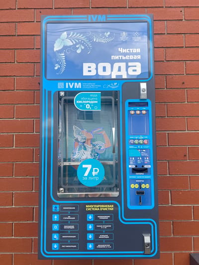 This image shows a water vending machine located outdoors, possibly in Mezhdurechenskiy, Russia, as indicated by the text on the machine. The machine is blue and grey, with a prominent display screen on the left showing text and a decorative floral design. The text on the screen reads "Чистая питьевая вода" which translates to "Pure drinking water". Below this, it says "IVM International Vending Machines". The right side of the machine has a coin slot, buttons for selecting volume, and a display showing the price. Text here indicates options for different coin denominations such as 1P, 2P, 5P, 10P, 50P, and 100P. There is also text describing the water purification system, including "ОЗОНИРОВАНИЕ" (Ozonation), "СТЕРИЛИЗАЦИЯ" (Sterilization), "ОБОГАЩЕНИЕ КИСЛОРОДОМ" (Oxygenation), "МИНЕРАЛИЗАЦИЯ" (Mineralization), and "ПОСТ-ФИЛЬТРАЦИЯ" (Post-filtration).
The machine is situated against a brick wall, suggesting it is in a public area. The lighting appears to be natural daylight, and the weather seems to be clear. The overall scene depicts a functional object in a typical urban or semi-urban setting. There are no people visible in the frame. The central part of the machine features a large transparent window through which a graphic illustration of a woman in traditional clothing can be seen. Above this graphic, the price for water is clearly displayed as "7 ₽ за литр", meaning "7 rubles per liter". The machine's design is modern and incorporates blue accents and stylized patterns.