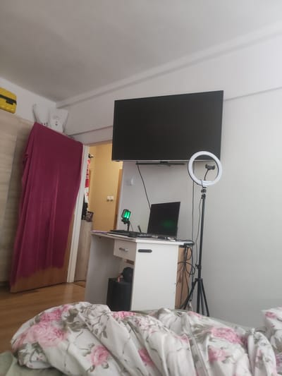 The image depicts a bedroom with a desk set up for content creation. A large, flat-screen television is mounted on the wall above a white desk. On the desk, there is a laptop, a keyboard, and a small green microphone. A ring light on a tripod stands next to the desk, suggesting the space is used for filming or streaming.

To the left of the desk, a wardrobe with a wooden finish is partially visible, with a bright yellow suitcase placed on top. A large, dark pink cloth hangs from a doorframe, obscuring the view of what lies beyond. The room appears to be tidy, with the focus on the technological setup.

In the foreground, a bed is covered with a floral duvet featuring pink roses and green leaves. The lighting suggests it is daytime, although the curtains are drawn, making it difficult to ascertain the exact time. The overall impression is that of a functional and personalized living space, likely in a residential setting, possibly Hradec Králové, Czechia. There is no visible text in the image.